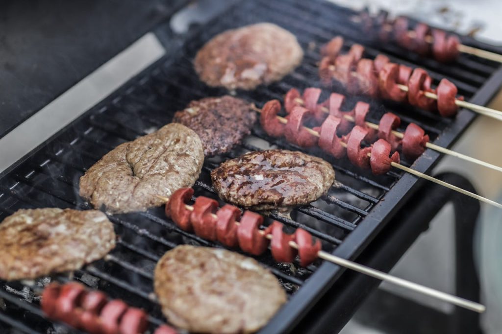 Close-up of grilled burgers and sausage skewers cooking on a barbecue grill outdoors.